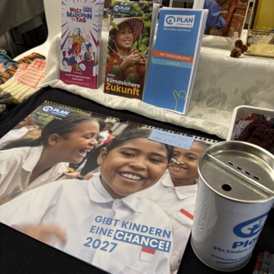 Stand von Plan International mit Informationsbroschüren, einem Kalender für das Jahr 2027 mit Bildern von Kindern und einer Spendendose.