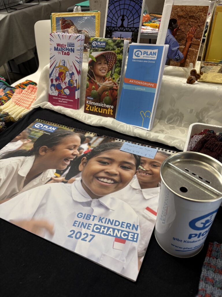 Stand von Plan International mit Informationsbroschüren, einem Kalender für das Jahr 2027 mit Bildern von Kindern und einer Spendendose.