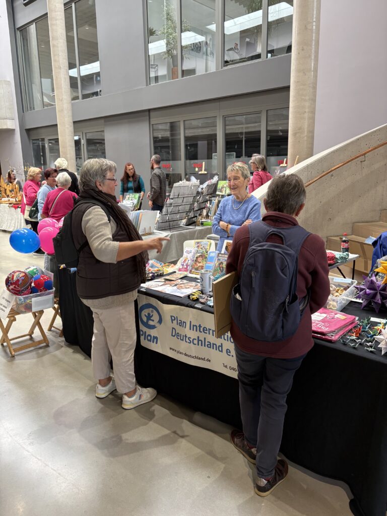 Zwei Frauen stehen an einem Stand von Plan International Deutschland auf einem Markt und unterhalten sich mit der Mitarbeiterin am Stand.