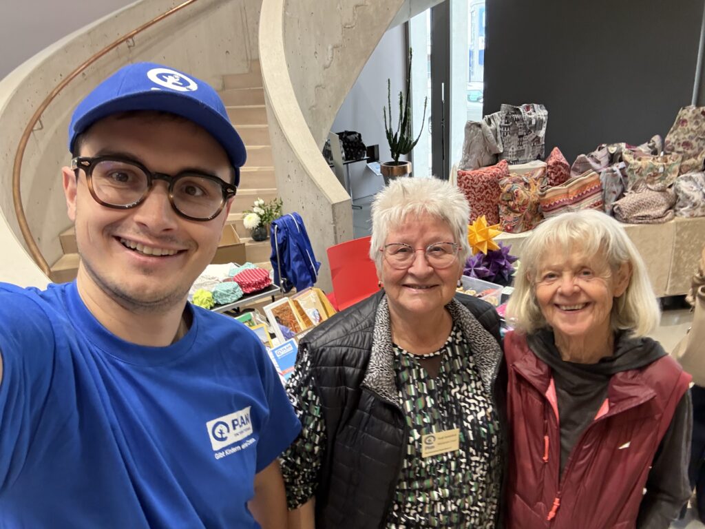 Ein junger Mann mit einer blauen Mütze und zwei ältere Frauen lächeln für ein Selfie vor einem Tisch mit verschiedenen Gegenständen und einer Treppe.